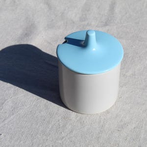 May include: A white ceramic sugar bowl with a light blue lid. The lid has a unique, rounded shape with a small spout.