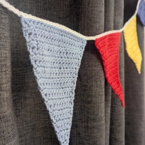 Op de afbeelding: Een handgemaakte gehaakte slinger met driehoekige vlaggen in lichtblauw, rood en geel. De vlaggen zijn aan elkaar geregen met een wit gehaakt koord, waardoor een decoratieve banner ontstaat. De slinger wordt getoond tegen een donkergrijze gordijn.