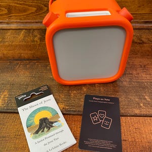 May include: An orange Yoto Player with a gray screen, a handle, and two side buttons. Beside it are two decks of cards. One card deck is labeled "The Shoot of Jesse" and the other says "Plays on Yoto".