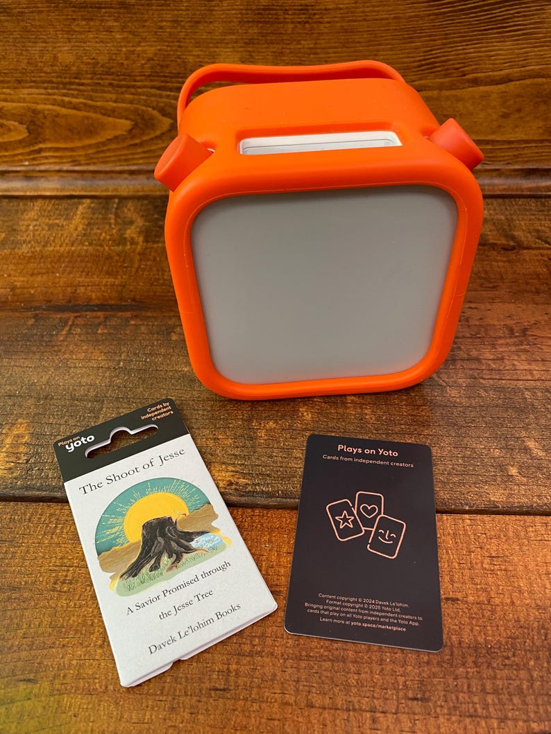 May include: An orange Yoto Player with a gray screen, a handle, and two side buttons. Beside it are two decks of cards. One card deck is labeled "The Shoot of Jesse" and the other says "Plays on Yoto".