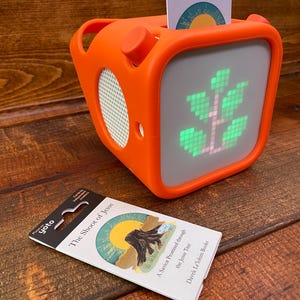 May include: An orange, cube-shaped speaker with a screen displaying a green tree graphic. A card labeled "The Shoot of Jesse" is inserted in the top. A second card with the same title and artwork lies in front of the speaker.