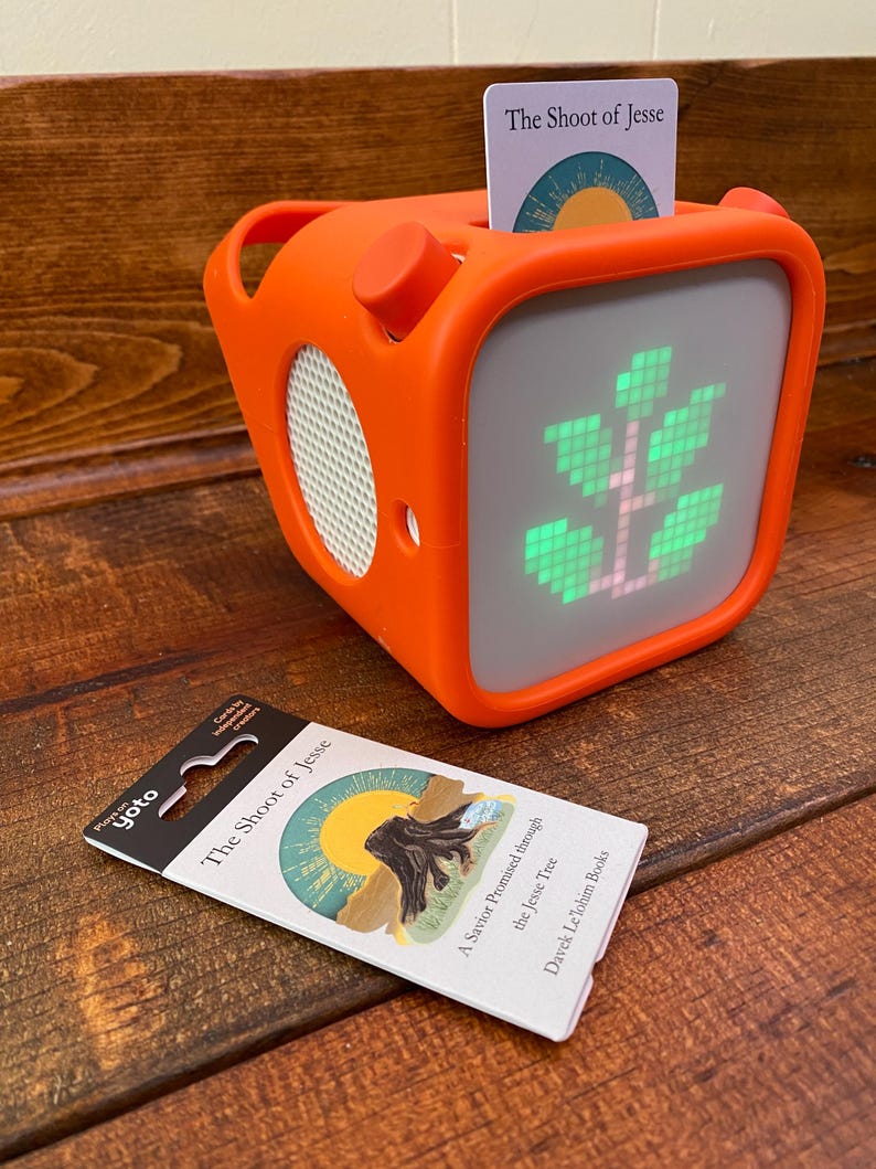 May include: An orange, cube-shaped speaker with a screen displaying a green tree graphic. A card labeled "The Shoot of Jesse" is inserted in the top. A second card with the same title and artwork lies in front of the speaker.
