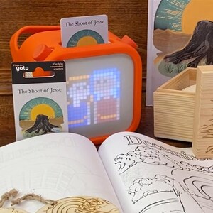 May include: An orange Yoto player with a lit screen displaying pixelated images, accompanied by a card set titled "The Shoot of Jesse." A wooden box and a book with illustrations are also present, suggesting a children's educational theme.