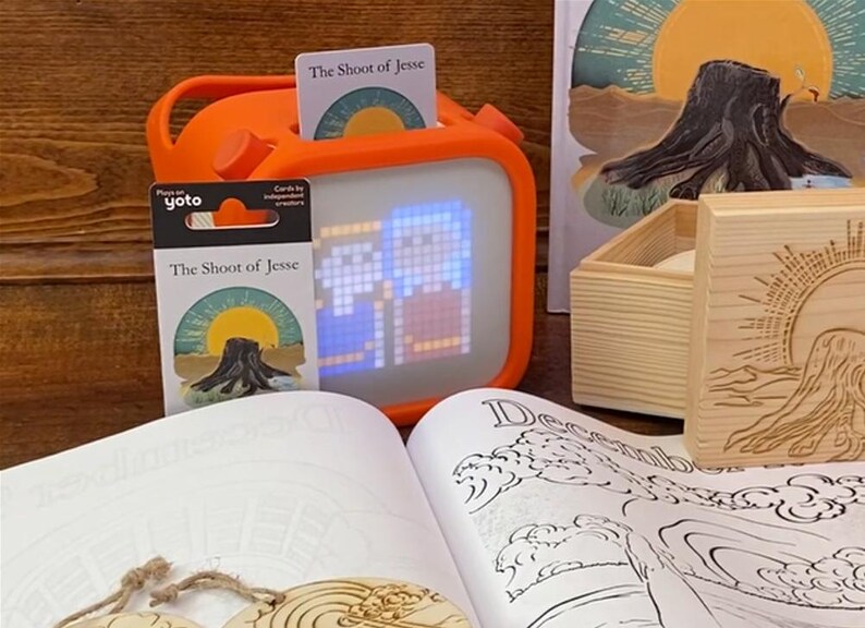 May include: An orange Yoto player with a lit screen displaying pixelated images, accompanied by a card set titled "The Shoot of Jesse." A wooden box and a book with illustrations are also present, suggesting a children's educational theme.