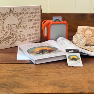 May include: Wooden plaque with etched text and a tree stump design, accompanied by a book, orange speaker, and wooden ornaments. The plaque reads "I AM THE ROOT AND SAPLING OF DAVID; THE STAR THAT SIGNALS THE BREAKING OF DAWN."