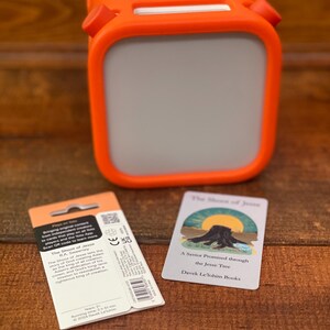 May include: An orange and gray portable speaker with a handle, a product package, and a card with the text "The Shoot of Jesse" and "A Savior Promised through the Jesse Tree" on a wooden surface.