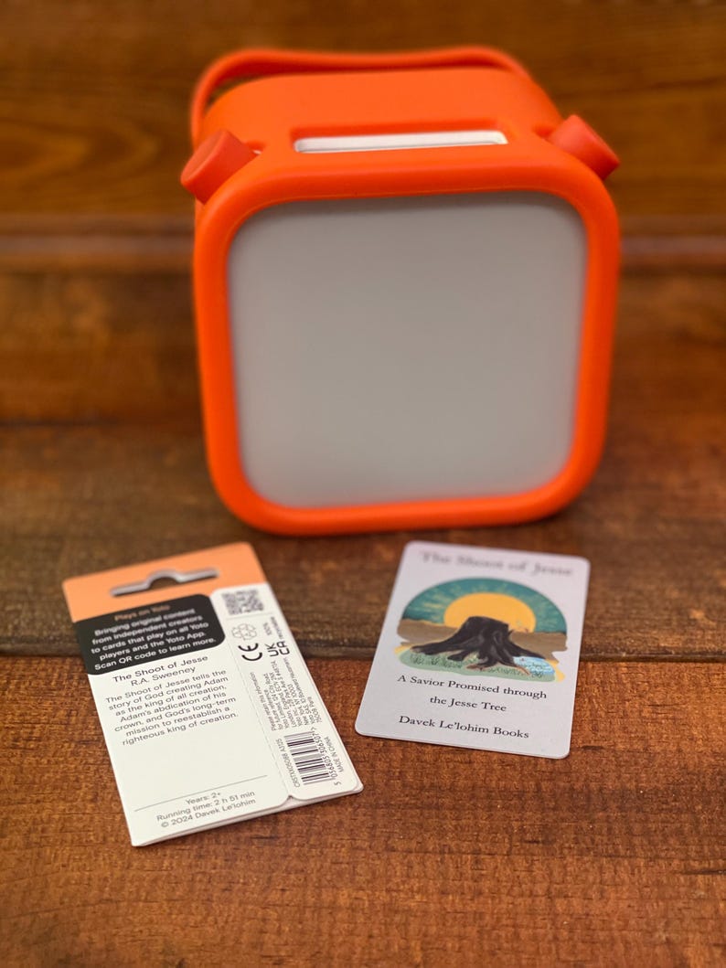 May include: An orange and gray portable speaker with a handle, a product package, and a card with the text "The Shoot of Jesse" and "A Savior Promised through the Jesse Tree" on a wooden surface.