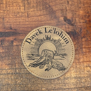 May include: A round, tan leather coaster with the text "Davek Le'lohim" encircling a central design. The design features a sun setting behind a tree stump and mountains. The coaster has a stitched edge.