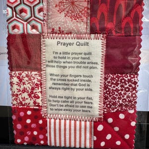 May include: A small red and white patchwork quilt with a message that reads "I'm a little prayer quilt to hold in your hand. I will help when trouble arises, those things you did not plan. When your fingers touch the cross tucked inside, remember that God is always right by your side. Hold me tight in your fist, to help calm all your fears. Don't be afraid to use me to wipe away your tears."
