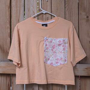 May include: A peach-coloured cropped t-shirt with a floral print pocket. The shirt has short sleeves and a crew neck.