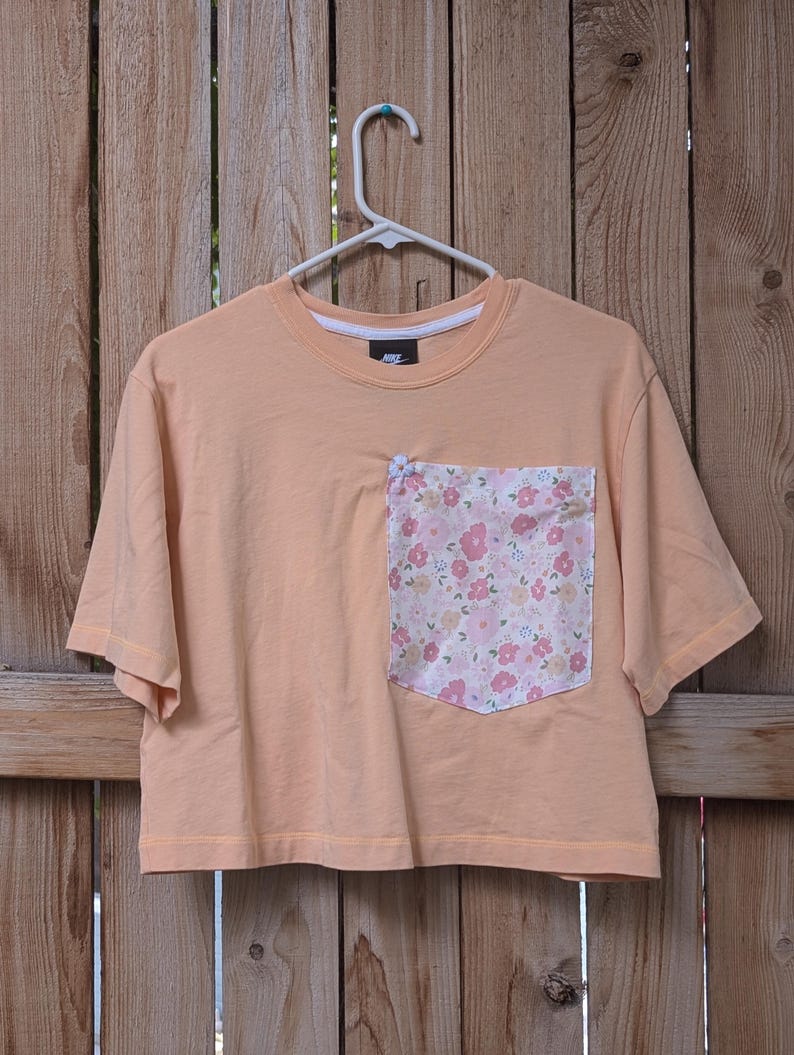 May include: A peach-coloured cropped t-shirt with a floral print pocket. The shirt has short sleeves and a crew neck.