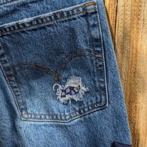 Pu&ograve; includere: Jeans di denim blu con una tasca posteriore che ha un piccolo cerotto floreale cucito sopra un buco. I jeans hanno un cerotto blu scuro nell'angolo in basso a destra.