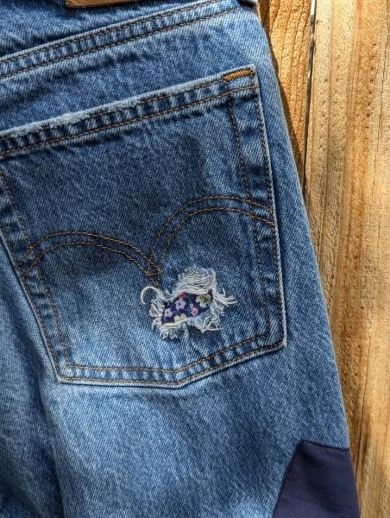 Pu&ograve; includere: Jeans di denim blu con una tasca posteriore che ha un piccolo cerotto floreale cucito sopra un buco. I jeans hanno un cerotto blu scuro nell'angolo in basso a destra.