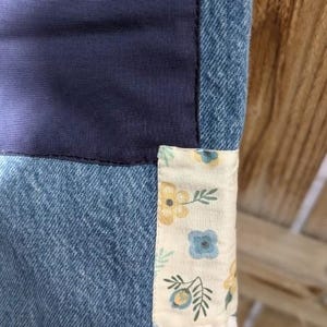 Pu&ograve; includere: Un primo piano di una trapunta di jeans con una toppa di tessuto floreale. La trapunta &egrave; realizzata in diverse tonalit&agrave; di denim blu e presenta uno sfondo bianco con fiori blu e gialli.