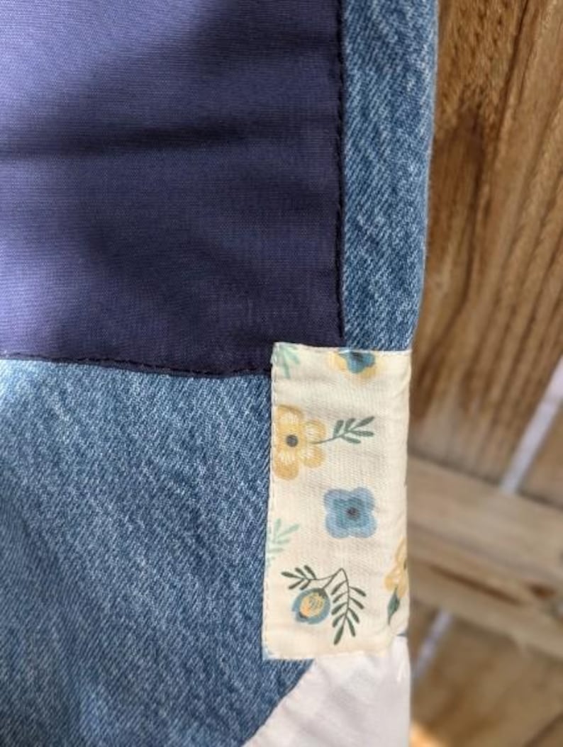 Pu&ograve; includere: Un primo piano di una trapunta di jeans con una toppa di tessuto floreale. La trapunta &egrave; realizzata in diverse tonalit&agrave; di denim blu e presenta uno sfondo bianco con fiori blu e gialli.