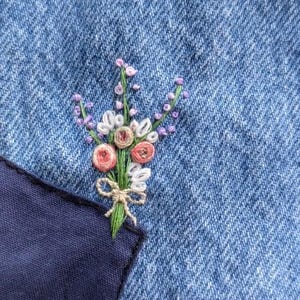 Pu&ograve; includere: Primo piano di un piccolo bouquet di fiori ricamati su un tessuto di jeans blu. I fiori sono rosa, bianchi e viola, e gli steli sono verdi. I fiori sono ricamati su una tasca del tessuto di jeans.