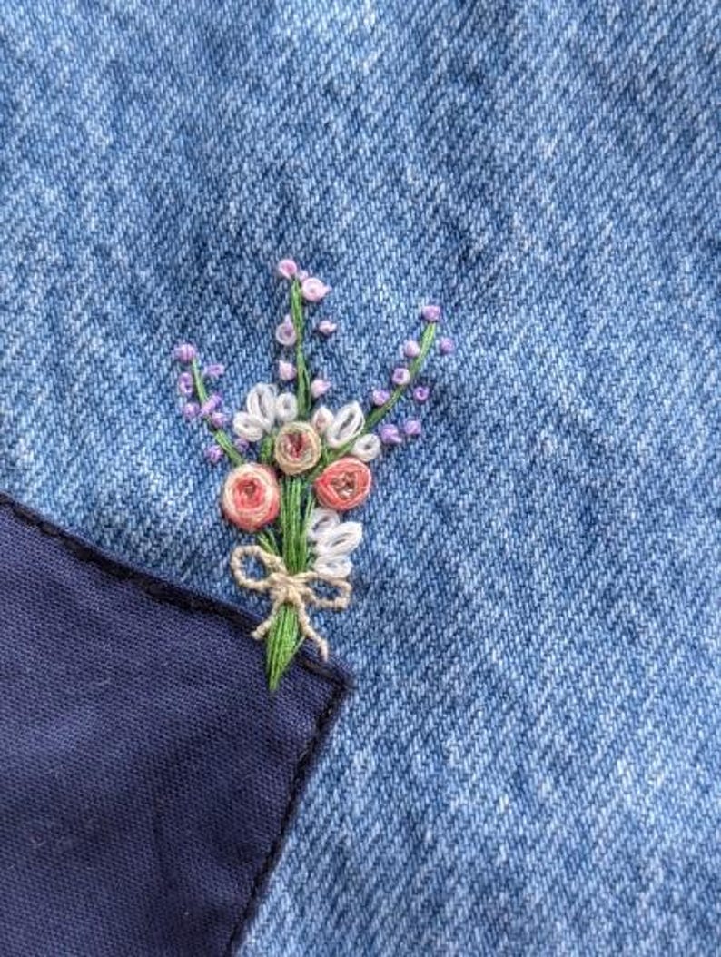 Pu&ograve; includere: Primo piano di un piccolo bouquet di fiori ricamati su un tessuto di jeans blu. I fiori sono rosa, bianchi e viola, e gli steli sono verdi. I fiori sono ricamati su una tasca del tessuto di jeans.