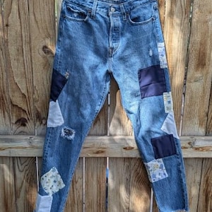 Pu&ograve; includere: Un paio di jeans blu denim con toppe di tessuti diversi cucite sopra. Le toppe sono in diverse tonalit&agrave; di blu, bianco e con motivi floreali.