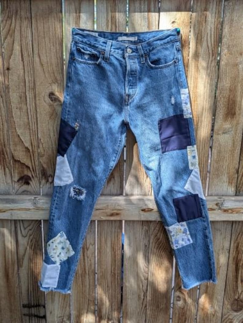 Pu&ograve; includere: Un paio di jeans blu denim con toppe di tessuti diversi cucite sopra. Le toppe sono in diverse tonalit&agrave; di blu, bianco e con motivi floreali.
