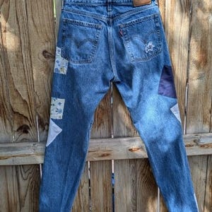 Pu&ograve; includere: Jeans di denim blu con toppe di tessuto in bianco, blu e nero. Le toppe sono cucite sul davanti dei jeans, sulle gambe e sulle tasche posteriori.