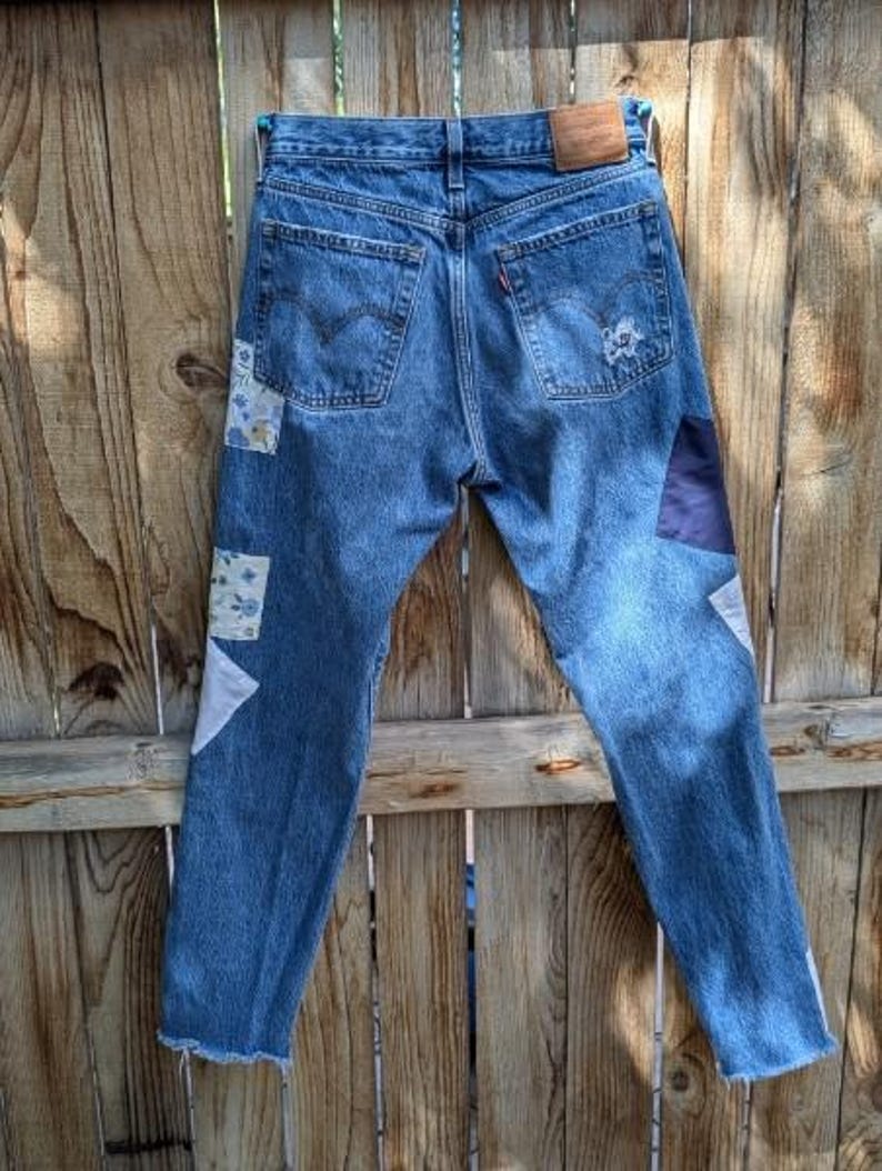 Pu&ograve; includere: Jeans di denim blu con toppe di tessuto in bianco, blu e nero. Le toppe sono cucite sul davanti dei jeans, sulle gambe e sulle tasche posteriori.