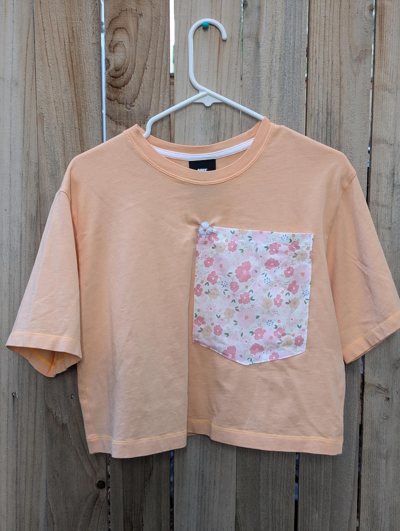 May include: A peach-coloured cropped t-shirt with a floral print pocket. The pocket has a white flower sewn on the top.