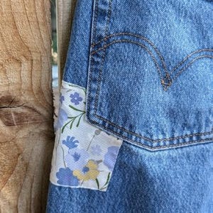 Pu&ograve; includere: Jeans blu denim con una toppa floreale cucita sulla tasca anteriore.