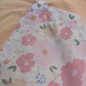 May include: A peach-coloured fabric with a white floral pattern. The fabric has a small white embroidered flower in the top corner.