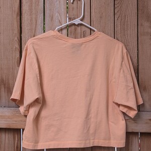 May include: A peach-coloured cropped t-shirt with short sleeves hanging on a white hanger against a wooden background.
