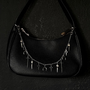 May include: A black hobo-style shoulder bag with a textured surface, featuring a silver-toned chain draped across the front. The chain is adorned with small white beads and metal charms shaped like crescent moons, four-pointed stars, and swords.