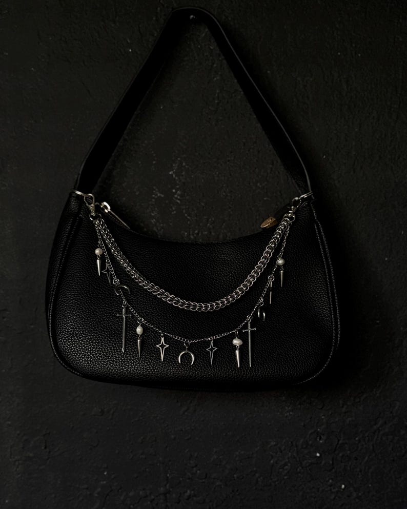 May include: A black leather handbag with a silver chain strap and multiple silver charms hanging from it. The charms include crosses, stars, and crescent moons.