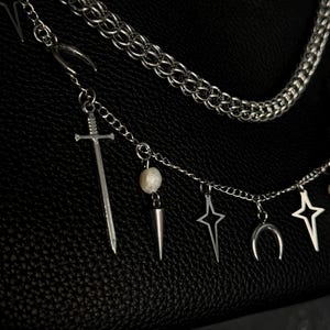 May include: A silver chain necklace with multiple charms, including a sword, a pearl, a star, a crescent moon, and a spike. The necklace is on a black leather background.