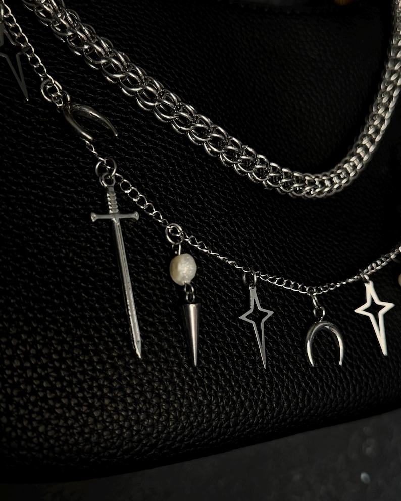 May include: A silver chain necklace with multiple charms, including a sword, a pearl, a star, a crescent moon, and a spike. The necklace is on a black leather background.