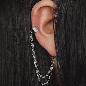 May include: Silver ear cuff with a chain design. The cuff wraps around the ear and connects to two dangling chains. The jewelry is worn on the ear, with black hair in the background.
