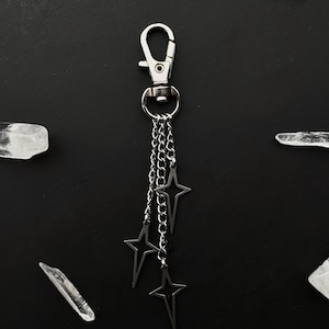 Puede incluir: Llavero plateado con tres colgantes de estrella colgando de delicadas cadenas. El llavero tiene un cierre de mosquetón para una fácil fijación. Las estrellas tienen un diseño minimalista y abierto. El fondo es negro liso, con fragmentos de cristal dispersos.