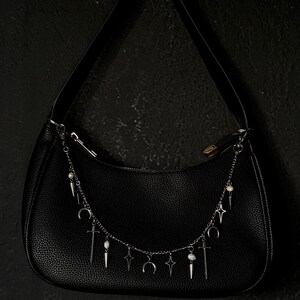 May include: A black leather handbag with a silver chain strap adorned with charms. The charms include crescent moons, stars, swords, and small white beads.
