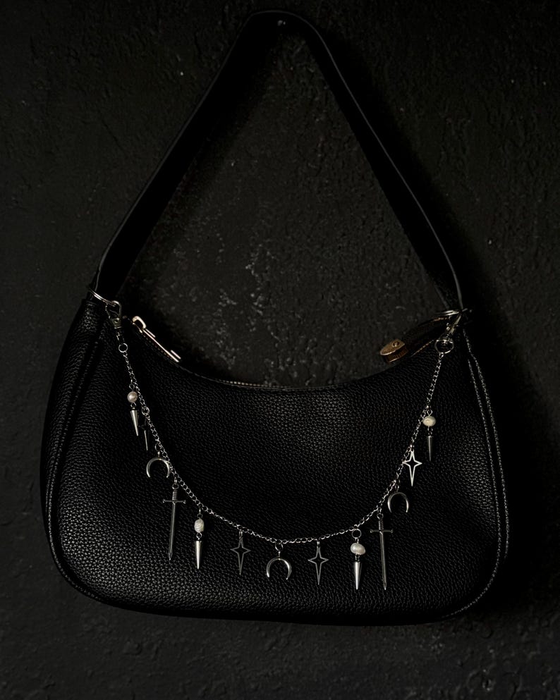 May include: A black leather handbag with a silver chain strap adorned with charms. The charms include crescent moons, stars, swords, and small white beads.