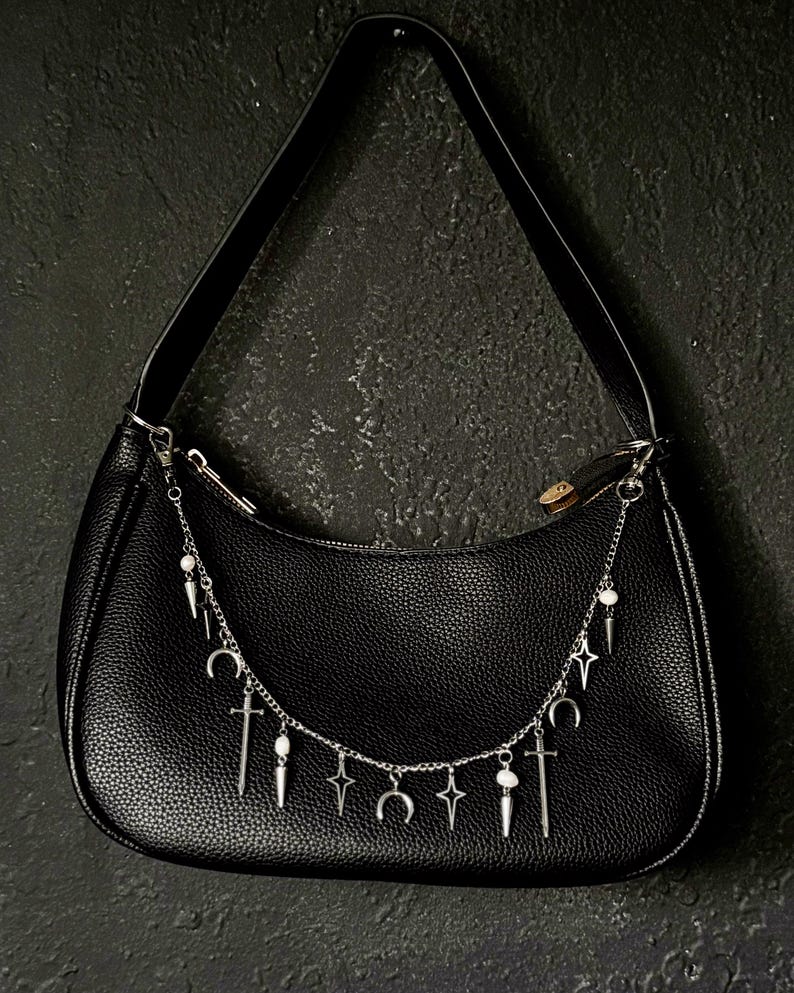 May include: Black leather handbag with a silver chain strap adorned with charms, including crescent moons, stars, and swords.