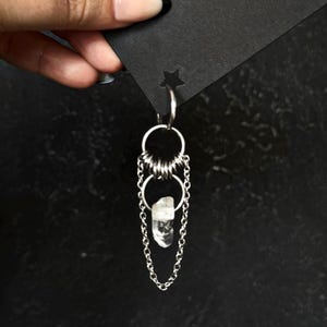 May include: A silver earring with a clear quartz crystal hanging from a chain with two silver hoops. The earring is hanging from a black card with a star cutout.
