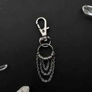 Puede incluir: Un llavero plateado con un dije circular y varias cadenas colgantes. El dije tiene un anillo de pequeños anillos de metal alrededor del centro.