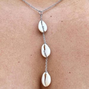 May include: A silver-toned necklace featuring three white cowrie shells suspended from a delicate chain. The shells are arranged in a vertical line, creating a simple yet elegant design. The necklace is a piece of jewelry.
