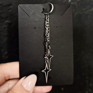 May include: A silver hoop earring with a dangling chain and three star-shaped charms. The earring is displayed on a black card.