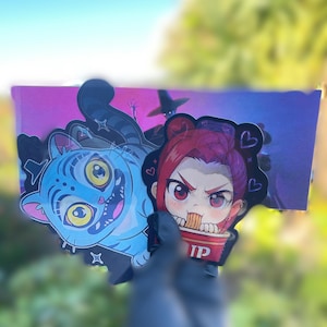 May include: Two cartoon stickers are held up against a blurred background. One sticker features a blue cat with yellow eyes and a striped tail. The other sticker depicts a red-haired anime character eating noodles from a cup with the letters "IP" on it.