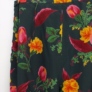 Puede incluir: Pantalones con estampado floral verde oscuro con un patr&oacute;n vibrante de flores y hojas amarillas, rosas y rojas. Los pantalones cuelgan de una percha plateada, mostrando el dise&ntilde;o de cintura alta y el intrincado patr&oacute;n floral.