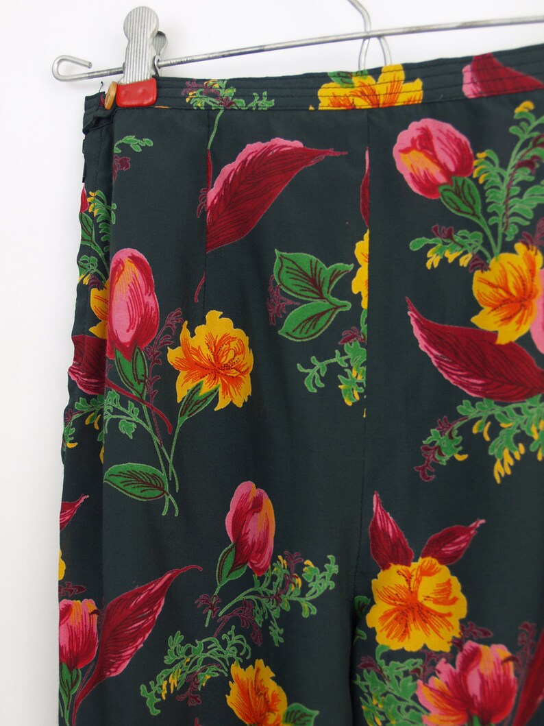 Puede incluir: Pantalones con estampado floral verde oscuro con un patr&oacute;n vibrante de flores y hojas amarillas, rosas y rojas. Los pantalones cuelgan de una percha plateada, mostrando el dise&ntilde;o de cintura alta y el intrincado patr&oacute;n floral.