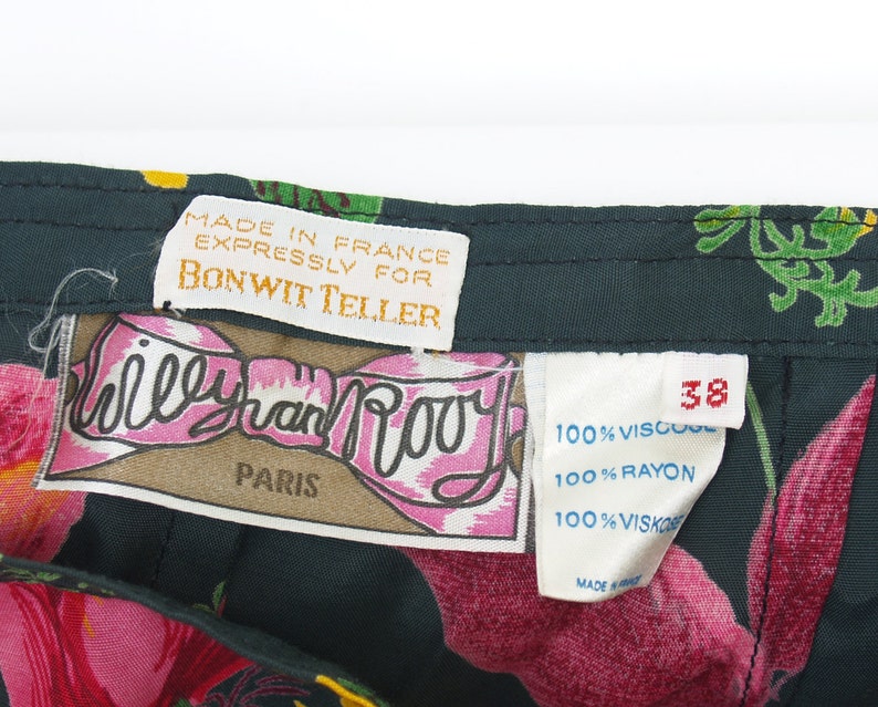 Puede incluir: Primer plano de una tela oscura con estampado floral con varias etiquetas. Las etiquetas dicen "MADE IN FRANCE EXPRESSLY FOR BONWIT TELLER" y "Willy van Rooy PARIS". La tela tiene un patr&oacute;n de grandes flores rosas y hojas verdes.