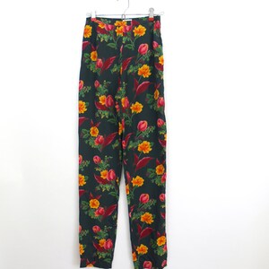 Puede incluir: Pantalones de cintura alta con un fondo verde oscuro y un vibrante estampado floral. El patr&oacute;n incluye flores y hojas amarillas y rojas. Los pantalones est&aacute;n colgados de una percha plateada sobre un fondo blanco.