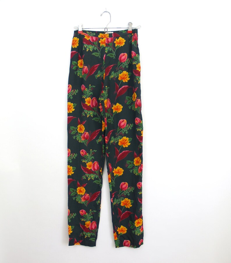 Puede incluir: Pantalones de cintura alta con un fondo verde oscuro y un vibrante estampado floral. El patr&oacute;n incluye flores y hojas amarillas y rojas. Los pantalones est&aacute;n colgados de una percha plateada sobre un fondo blanco.
