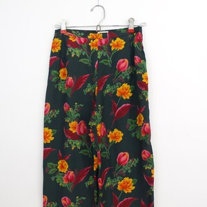 Puede incluir: Pantalones con estampado floral verde oscuro colgados de una percha met&aacute;lica. Los pantalones presentan un patr&oacute;n repetido de flores amarillas y rojas con hojas verdes. Los pantalones son de tejido ligero.