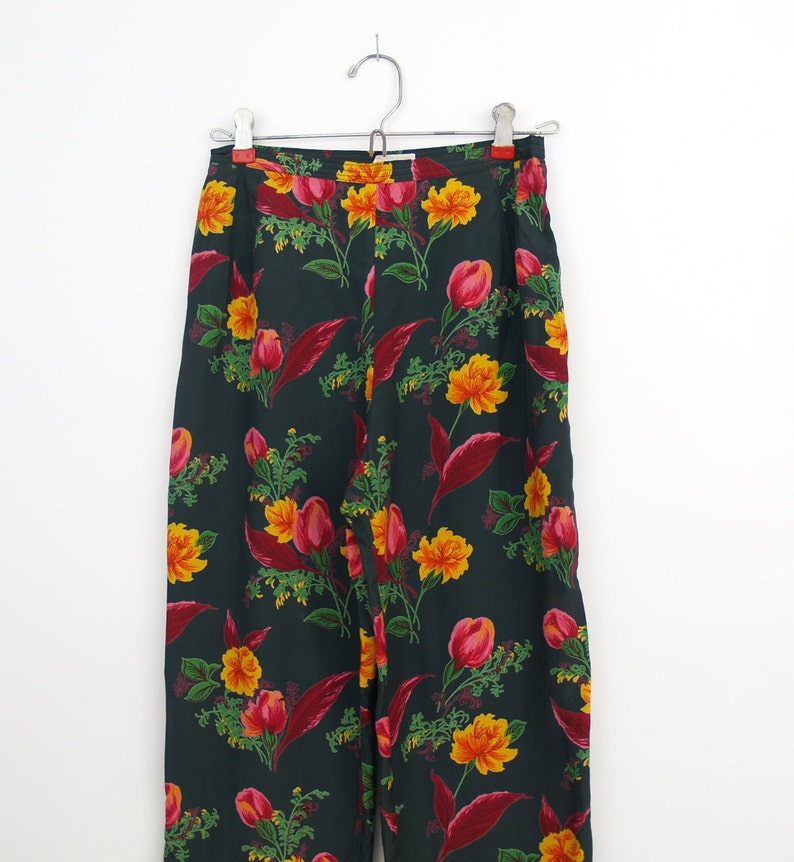 Puede incluir: Pantalones con estampado floral verde oscuro colgados de una percha met&aacute;lica. Los pantalones presentan un patr&oacute;n repetido de flores amarillas y rojas con hojas verdes. Los pantalones son de tejido ligero.